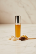 The Signal Roller — Scent-Memory Anchor. Frosted glass roller bottle with amber-gold liquid and silver cap on cream marble, surrounded by frankincense resin and tonka bean. Warm editorial wellness aesthetic.